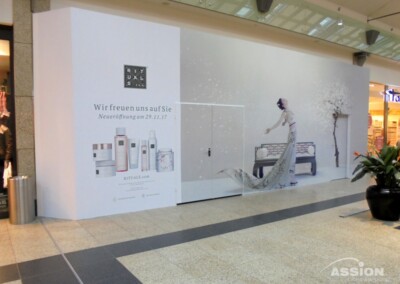 Eine Schaufensterfront mit einer Rituals-Werbung, die die Eröffnung am 29. November 2017 ankündigt, mit Produktbildern und einer Frau, die nach einer Bank greift.