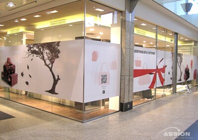 Schaufenster aus Glas mit großen Aufklebern, die einen Baum, fallende Blätter, Geschenkboxen, Handtaschen, eine Schleife und einen QR-Code zeigen. Das Ladeninnere ist durch die Fenster sichtbar.