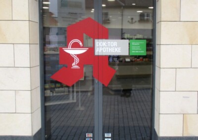 Gläserne Eingangstür einer Apotheke mit einem großen roten Apothekensymbol und der Aufschrift "Doktor Apotheke"; im Glas sind Spiegelungen von Gebäuden zu sehen.