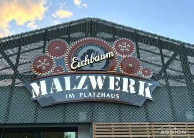 Schild für "Eichbaum Malzwerk im Platzhaus" mit dekorativen Zahnrädern auf einer Metallgitterfassade, bei Tageslicht fotografiert.