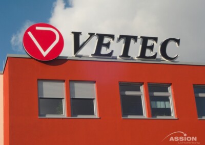Ein rotes Gebäude mit drei Fenstern und einem großen Schild auf dem Dach mit der Aufschrift "VETEC" und einem roten kreisförmigen Logo links neben dem Text.