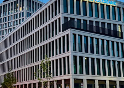 Modernes Bürogebäude mit dem Schild "Heidelberger Volksbank" auf dem Dach, mit großen Fenstern, mehreren Stockwerken und einigen kleinen Bäumen davor.