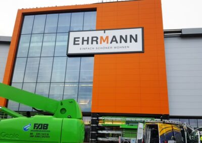 Außenansicht einer Ehrmann-Filiale mit orangefarbener Fassade, großen Glasfenstern und vor dem Gebäude sichtbaren Baumaschinen.