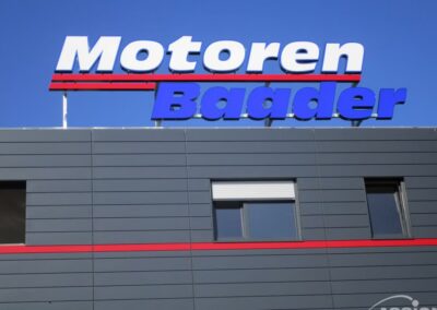 Auf der Außenseite eines grauen Gebäudes ist das Schild "Motoren Baader" in großen blauen und weißen Buchstaben vor einem klaren blauen Himmel zu sehen.