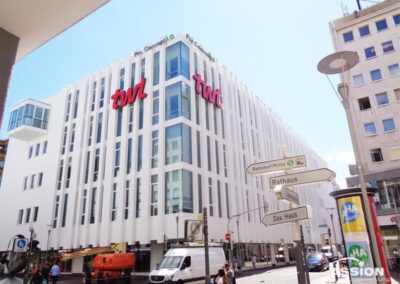 Großes weißes modernes Bürogebäude mit "twi"-Schildern, Straßenschildern im Vordergrund, Fahrzeugen und Fußgängern auf der Straße und klarem Himmel darüber.