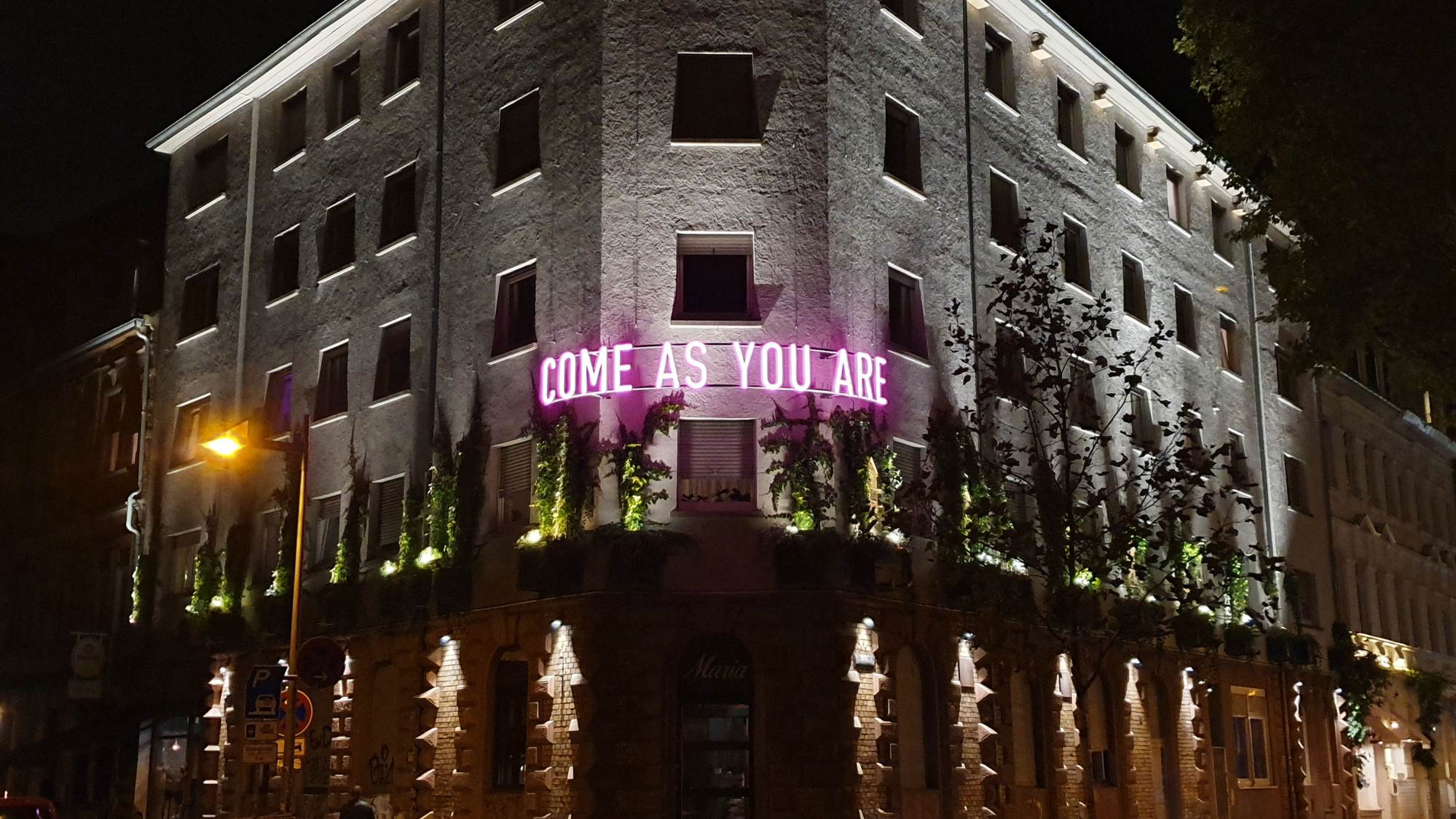 Ein Gebäude bei Nacht mit beleuchteten Fenstern und einem rosafarbenen Neonschild mit der Aufschrift "COME AS YOU ARE" über dem Erdgeschoss, umgeben von etwas Grün.