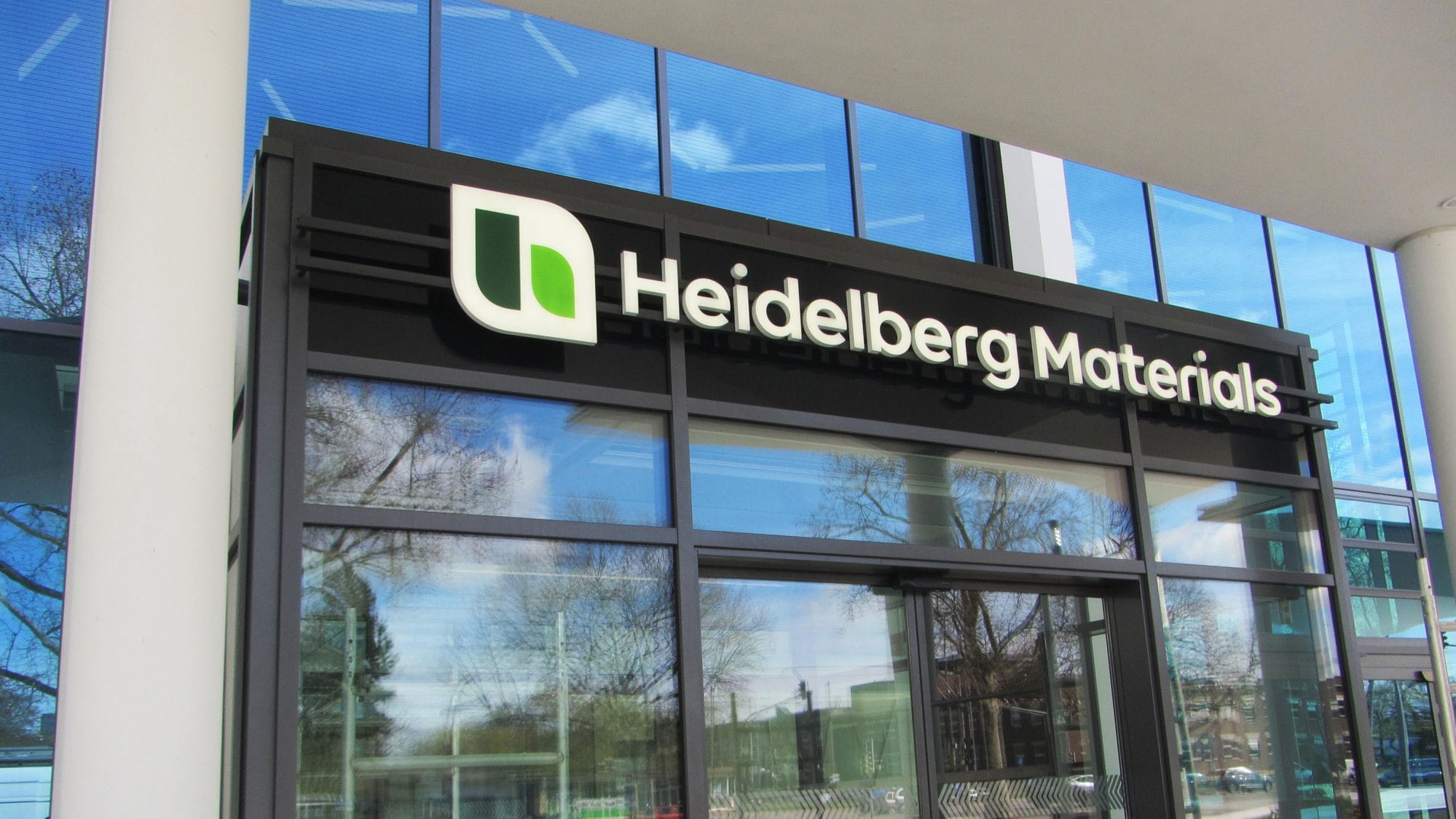 Der Eingang eines modernen Gebäudes mit einer Glasfassade trägt das Logo und den Namen "Heidelberg Materials" über den Türen.