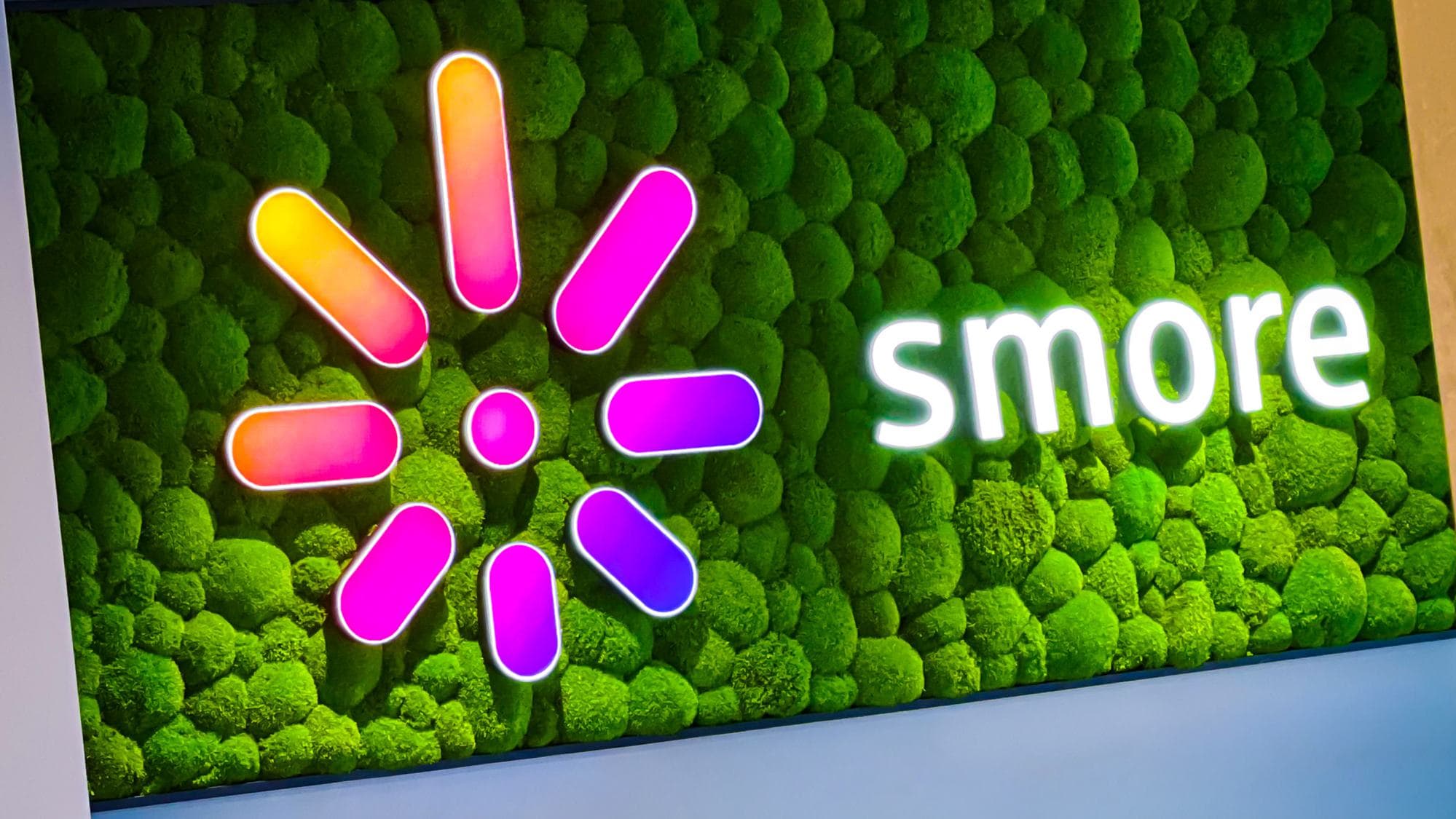 Beleuchtetes "Smore"-Logo mit einem farbenfrohen radialen Muster auf einem grünen Mooswand-Hintergrund.