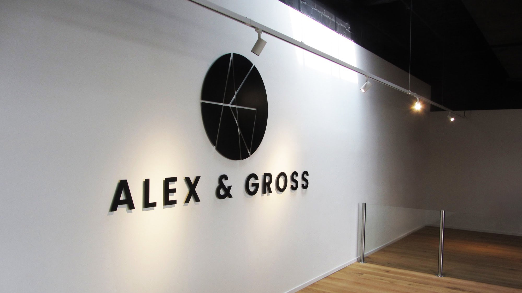 Weiße Wand mit schwarzem, kreisförmigem Logo und dem Text "ALEX & GROSS" in großen Buchstaben; Holzboden und Strahler an der Decke.