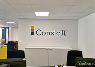 Eine moderne Büroeinrichtung mit Innenwerbung mit dem "Constaff"-Logo an einer weißen Wand über einem Schrank, Schreibtischen mit Stühlen im Vordergrund und Fenstern auf der linken Seite.