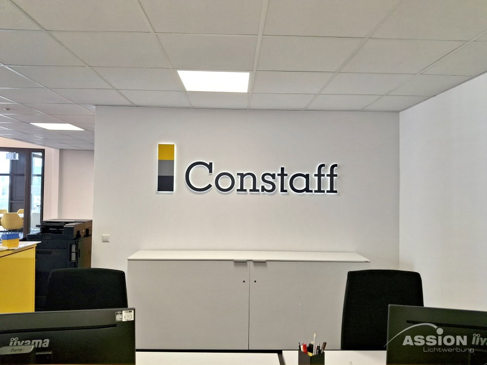 Innenwerbung (1) XXX Eine moderne Büroeinrichtung mit Innenwerbung mit dem "Constaff"-Logo an einer weißen Wand über einem Schrank, Schreibtischen mit Stühlen im Vordergrund und Fenstern auf der linken Seite.