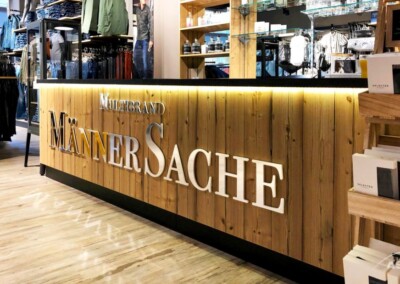 Holztheke mit dem Schild "Multibrand Männersache" in einem modernen Herrenbekleidungsgeschäft; im Hintergrund sind Regale mit Kleidung und verpackten Produkten zu sehen.