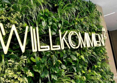 Beleuchtetes "Willkommen"-Schild an einer grünen Wand mit verschiedenen Pflanzen in einem Innenbereich.