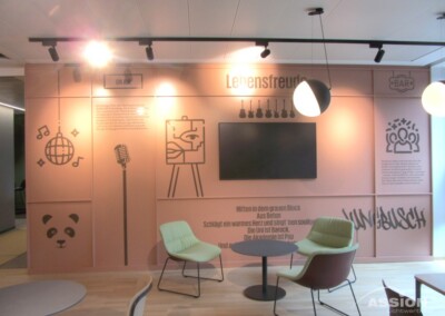 Moderner Lounge-Bereich mit pastellrosa Wand, montiertem Fernseher, minimalistischen Stühlen und gemalten Symbolen wie einem Panda, einer Discokugel, einem Mikrofon und einem Herz. An der Wand ist ein deutscher Text angebracht.