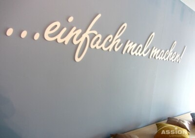 In weißer Schrift auf einer blauen Wand steht "...einfach mal machen!". Über einer Bank mit einigen Kissen; das Logo der ASSION Lichtwerbung befindet sich in der unteren rechten Ecke.