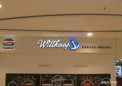 Ein Schild über einem Restaurant zeigt ein Burger-Symbol, den Namen "Wittkoop's" mit einem Anker und die Aufschrift "BØRGER HØKERS" in blauen Buchstaben.
