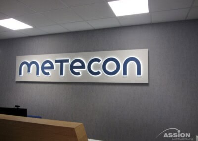 Innenraum eines Büros mit einem beleuchteten Schild mit dem Wort "metecon" an einer grauen Wand über einem hölzernen Empfangstresen.