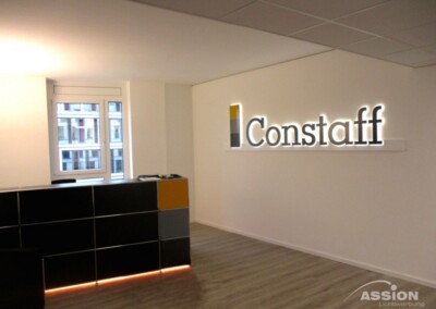 Moderner Büro-Empfangsbereich mit einem schwarz-gelben Schreibtisch und dem beleuchteten Constaff-Logo an einer weißen Wand. Im Hintergrund ist ein Fenster zu sehen.