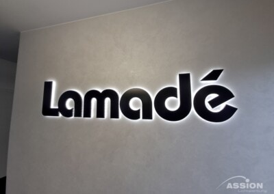 Schwarze 3D-Buchstaben mit dem Schriftzug "Lamadé" sind auf einer hinterleuchteten Lichtwand montiert; in der rechten unteren Ecke befindet sich das Logo "ASSION Lichtwerbung".