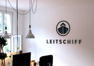 Modernes Büro mit einem Schreibtisch, einem Computer, einem Bürostuhl, Hängelampen und einem Wandlogo mit der Aufschrift "LEITSCHIFF". Der Raum ist minimalistisch mit natürlichem Licht durch ein großes Fenster.