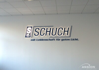 Das Bild zeigt ein Wandschild mit der Aufschrift "SCHUCH mit Leidenschaft für gutes Licht". In der rechten unteren Ecke ist das Logo der "Assion Lichtwerbung" zu sehen.