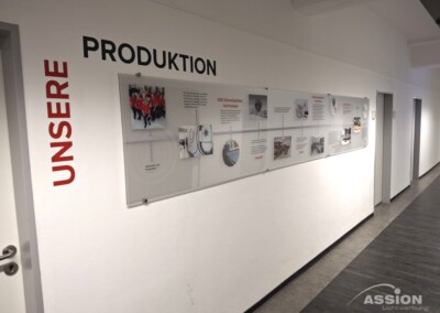 Wanddisplay im Flur mit dem Schriftzug UNSERE PRODUKTION" und einer Zeitleiste, die Produktionsprozesse und Bilder zeigt, in einem modernen Büro- oder Industriegebäude.