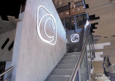 Eine moderne Treppe mit Glasgeländer, Betonwänden und einem großen beleuchteten "C"-Symbol an der Wand, die zu einer oberen Etage mit Holzakzenten führt.