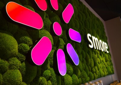 Ein leuchtendes Logo in Form einer rosa und lila Blume und das Wort "Smore" sind auf einer grünen Mooswand über einem Regal mit Flaschen zu sehen.