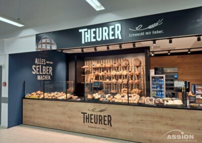Bäckertheke mit verschiedenen Brotsorten und Gebäck; an der Wand dahinter hängen hölzerne Utensilien; auf den Schildern steht "THEURER" und "Alles selbst machen".