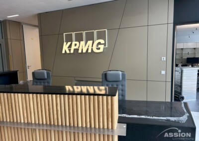 Empfangsbereich mit zwei Stühlen und einem Schreibtisch vor einer Wand mit dem beleuchteten KPMG-Logo; im Hintergrund ist das Büromobiliar zu sehen.