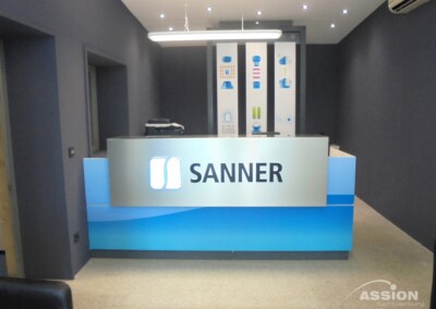 Empfangstresen mit "SANNER"-Logo in einem modernen Bürobereich mit blauen Akzenten, dunkelgrauen Wänden und Deckenstrahlern.