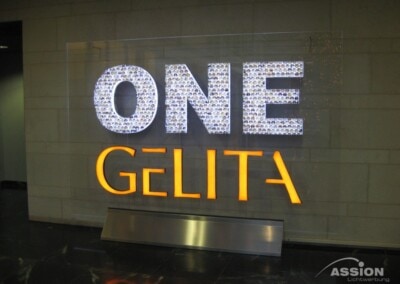 Beleuchtetes Wandschild mit der Aufschrift "ONE GELITA" in weißer und orangefarbener Schrift, montiert an einer Steinwand über einem dunklen Marmorboden.