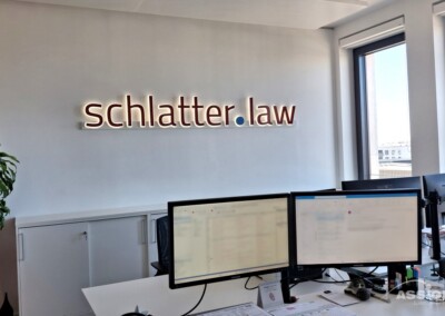 Modernes Büro mit zwei Computermonitoren auf einem Schreibtisch, einer Pflanze in der Ecke und einem Wandschild mit der Aufschrift "schlatter.law" in Leuchtbuchstaben.