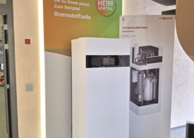 Ein weißes Brennstoffzellen-Heizgerät steht vor einer Informationswand mit deutschem Text und einem technischen Diagramm auf einer Indoor-Ausstellung.