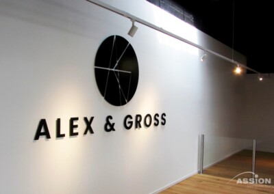 Eine weiße Wand mit einem schwarzen geometrischen Logo über dem Schriftzug "ALEX & GROSS" in fetten Buchstaben; Parkettboden und Schienenbeleuchtung sind sichtbar.