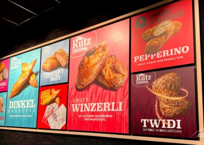 An der Wand hängen bunte Plakate, auf denen verschiedene Brotprodukte der Marke Rütz, darunter Baguettes, Brote und Brötchen, mit deutschen Texten und Produktbildern beworben werden.