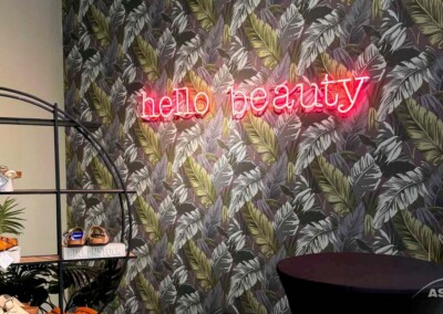 Ein pinkes Neonschild mit der Aufschrift "Hallo Schönheit" ist an einer mit Blättern tapezierten Wand neben einer Auslage mit Schuhen und einem schwarzen runden Tisch angebracht.