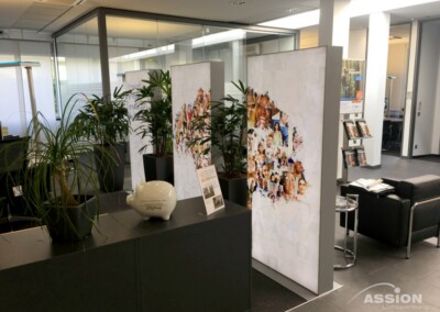 Moderne Bürolobby mit schwarzen Sitzmöbeln, Zimmerpflanzen, Prospektständern, einem Sparschwein auf einem Tresen und beleuchteten Schautafeln mit einer Collage aus Gesichtern.