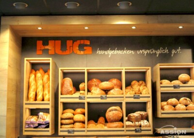 Regale in einer Bäckerei mit verschiedenen Brotsorten und Baguettes unter einem Schild mit der Aufschrift "HUG handgebacken. ursprünglich. gut.