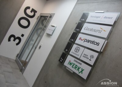 Hinweisschild mit Firmennamen und -logos an einer Betonwand neben einer Glastür im dritten Stock eines Gebäudes.