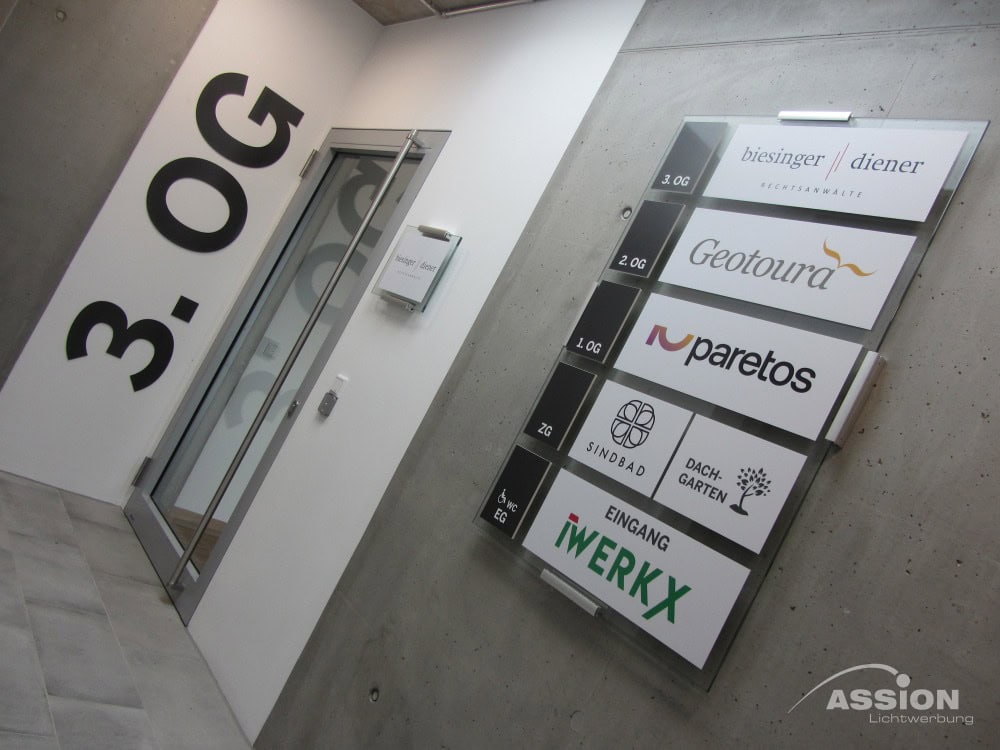 Hinweisschild mit Firmennamen und -logos an einer Betonwand neben einer Glastür im dritten Stock eines Gebäudes.