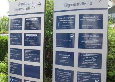 Zwei große Wegweiser zeigen die Namen und Fachgebiete der Arztpraxen im Ärztehaus 1 und 2, Hilgardstraße 26 und 30, mit einem Pfeil, der nach rechts zeigt.