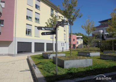 An einem sonnigen Tag steht zwischen modernen Wohngebäuden ein Innenhof mit erhöhten Pflanzgefäßen, kleinen Bäumen und einem Mast mit Wegweisern.
