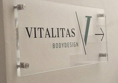 Ein durchsichtiges Wandschild mit der Aufschrift "Vitalitas Bodydesign" und einem Pfeil nach rechts ist an einer weißen Wand angebracht. Rechts unten ist das Logo von "Assion Lichtwerbung" zu sehen.