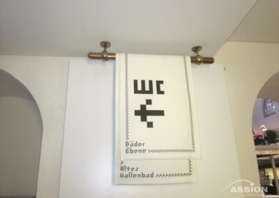Weißes Tuch mit einem schwarzen WC-Symbol im Pixel-Stil und deutschem Text hängt an einer Messingstange, die an einer weißen Wand befestigt ist.