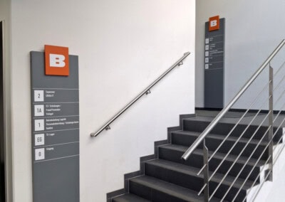 Innentreppe mit schwarzen Stufen, einem Metallhandlauf und an der Wand befestigten Wegweisern mit einem weißen "B" auf orangefarbenem Hintergrund. Helles, sauberes, modernes Interieur.