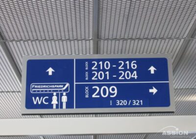 Blaues Schild im Stil eines Flughafens mit Hinweisen zu den Sitzplatzblöcken 210-216, 201-204 und 209 sowie zu den Parkplätzen (Friedrichspark) und den Toiletten (WC) mit Symbolen für Männer und Frauen.