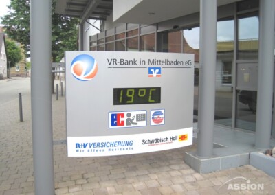 Ein digitales Schild vor der VR-Bank in Mittelbaden zeigt die Temperatur von 19°C an; darunter erscheinen Logos von Versicherungen und anderen Partnern.