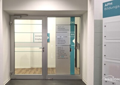 Glastür und Wand mit Beschilderung, die Raumrichtungen und Etagen in einem modernen Büro- oder Bildungsgebäude mit Holzboden und weißen Wänden anzeigt.