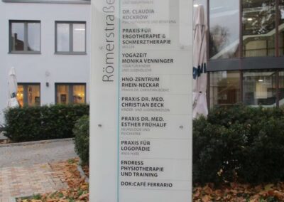 Ein Wegweiser in der Römerstraße listet mehrere Arzt- und Therapiepraxen auf, darunter Ärzte, Physiotherapeuten und ein Café, während in der Nähe Herbstlaub auf dem Boden liegt.